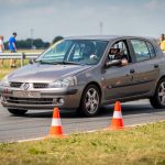 Comment débuter le sport automobile avec un budget limité ? Jeune homme amateur casqué au volant d'une petite citadine traversant un parcours de slalom bordé de cônes orange lors d'une journée sportive locale.