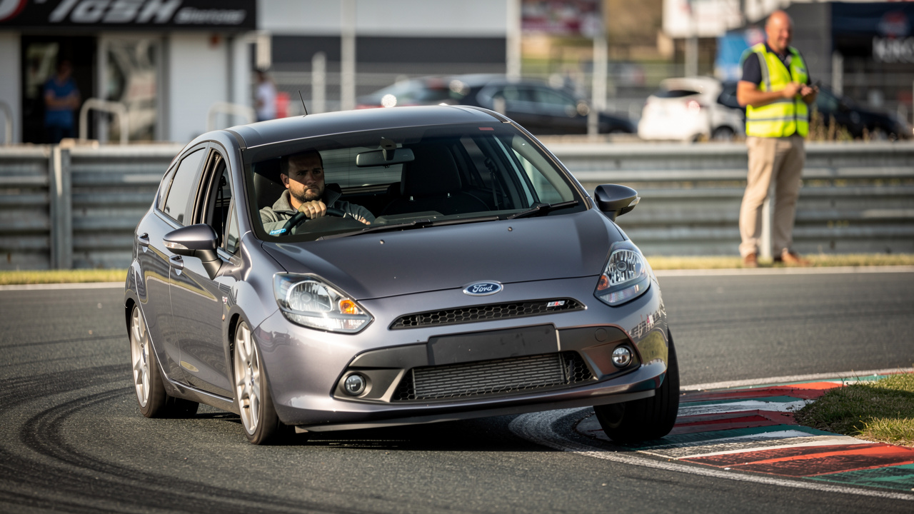 Voiture compacte de série négociant un virage sur un circuit, avec un pilote concentré, un commissaire de piste et un paddock sobre en arrière-plan.