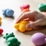 Quels critères vérifier avant d’acheter un squishy anti-stress ? Gros plan de plusieurs squishies anti-stress colorés de formes variées sur une table claire, tandis qu’une main de femme en presse doucement un.